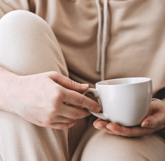 Person in loungewear holding a white mug while sitting with knees up on a sofa