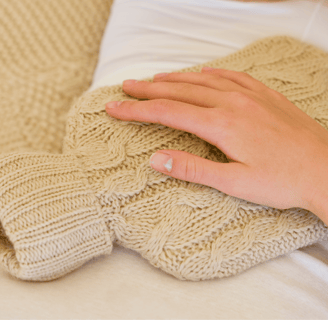 Person using a knitted hot water bottle on their lower abdomen, wearing cozy loungewear.