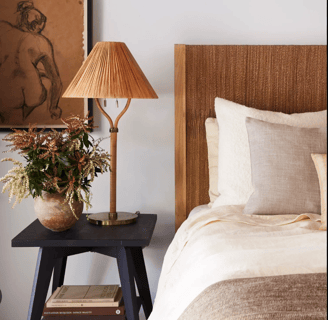 brown lamp on a black table beside cream color bedsheets with a neutral brown bed frame, plant