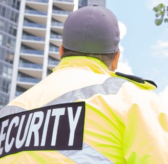 Ein Sicherheitsmitarbeiter mit einer "SECURITY"- Jacke mit dem Blick auf ein Hochhaus.