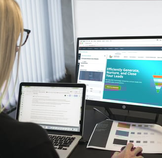 a woman sitting at a desk with a laptop and a monitor showing an email marketing platform