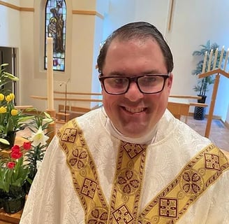Person in religious vestments standing in a church with flowers and stained glass window in the back