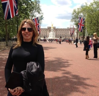 mulher loira, com óculos de sol, em frente a um palácio em rua com bandeiras britânicas