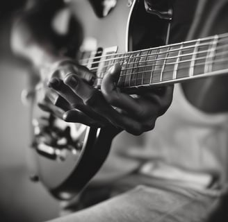 A person is playing an electric guitar. The image is captured in black and white, highlighting the musician's fingers on the fretboard and the guitar headstock with the brand logo visible. The focus is on the hand's positioning and the strings of the guitar.