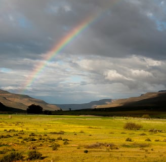 Rainbow across a cloudy sky with some fields in the foreground and a circular mountain range as backdrop