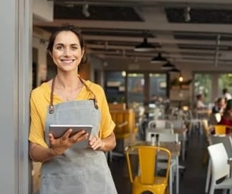 Uma empreendedora posando sorrindo na porta do seu restaurante, com um laptop nas mãos.