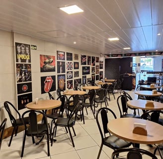 Área da cafeteria com mesas cadeiras e pôsteres de rock no Studio Brothers em Porto Alegre
