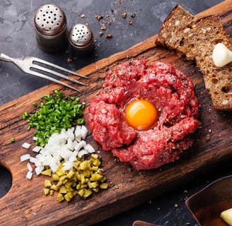 beef tartare on a wooden board, garnished with a raw egg yolk, diced onions, chives, pickles, rye bread, and a garlic clove