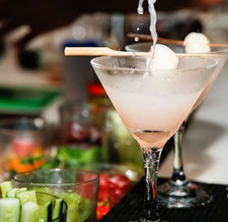 Refreshing lychee martini being poured into a cocktail glass with garnished lychees, set against a vibrant bar background