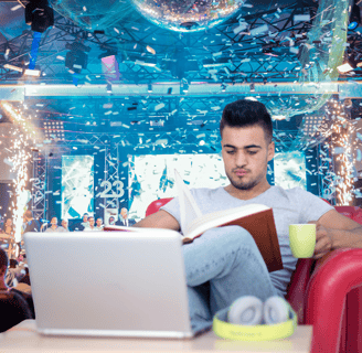 A man sitting in a red armchair holding a mug and reading a book with a party in the background