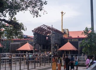 A temple entrance build in red color rocks with people standing in front of the temple.