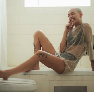 A woman sitting in the bathroom, on the lip on her bathtub, smiling and touching her face