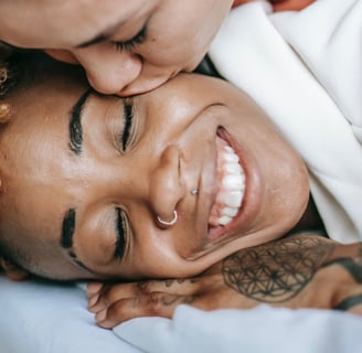 A woman kisses another woman's face while laying on a bed. The woman being kissed is smiling broadly