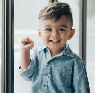 An adorable young boy standing in front of a window with his hand raised