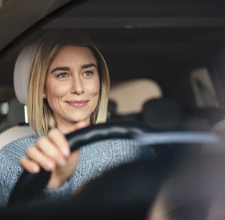 A person is seated in the driver's seat of a car, holding the steering wheel with one hand.