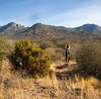 a man walking through a grassy area with mountains in the background