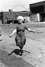 B&W image of elderly woman on a town back street. She is skipping with a skipping rope