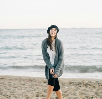 Woman smiling and standing on a beach wearing a sweater and beret.
