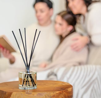 a group of women sitting on a couch reading a book and they sit behind a bottle of home scent