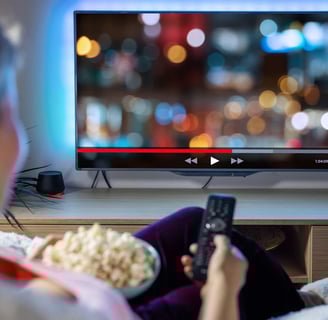 Uma pessoal segurando um balde de pipocas e com um controle remoto na mão assistindo a TV