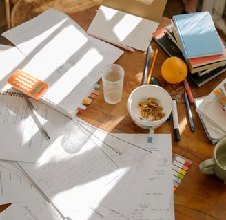 A cluttered round table, containing paper, crockery, food, books and stationary.