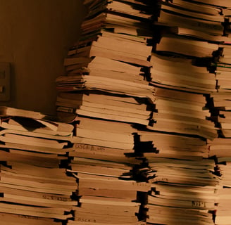 Multiple piles of books, stacked agains a wall. A pair of lightswitches is visible beside the piles.