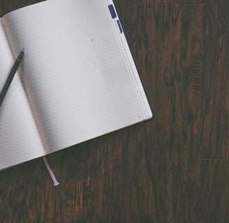 An open lined notebook on a wooden table, with a pen laid across it.