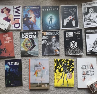 A selection of TTRPGs, mostly zines, laid out on a carpet in rows.