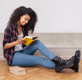 A young woman seated on the floor, reading a book from a physical and digital magazine publisher