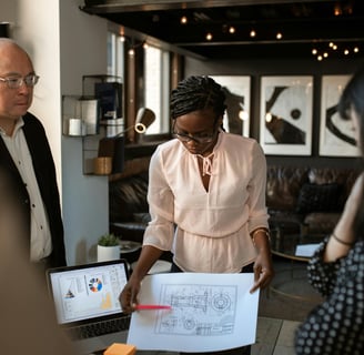 One man and two women standing at a table discussing business blue print schematics with a laptop displaying charts