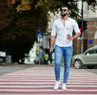 homem atravessa a rua em uma faixa de pedestres. Ele usa uma camisa branca, jeans e tênis branco