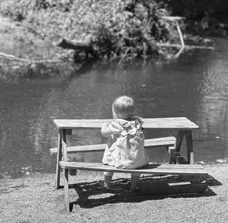 Black and white photo of a baby girl sitting at a miniture picnic table, gazing at a lake with trees in the distance. 