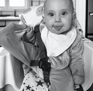 Black and white photo of smiling baby in high chair with tongue out, showing natural baby joy, innocence, and purity.