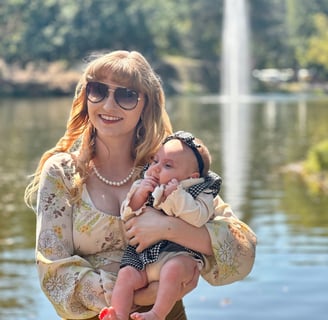 A mother cradling her newborn daughter in front of a lake, with a fountain softly spraying in the background.