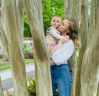 A mother holding her baby in a tree, kissing her on the cheek while the baby looks at the camera, showing loving mother-child