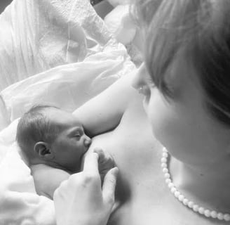 Black and white top view of new mom nursing baby in bed. Baby holds mother's finger as she gazes down with love.