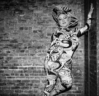 black & white drag queen posed fashionably in front of brick wall on stage in a patterned bodysuit