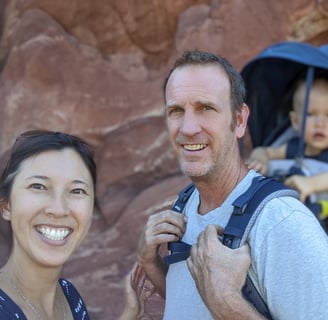Smiling couple hiking with their baby in a carrier against red rock cliffs in Colorado.