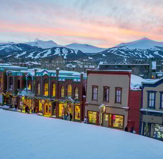 Snow-covered street lined with colorful historic buildings at sunset with ski slopes behind.