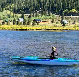 Young boy paddling a bright blue kayak on a calm river with forest and cabins in the background.