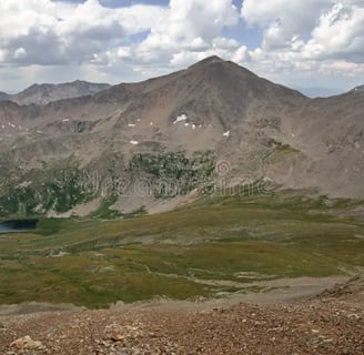 Expansive view of a rugged mountain range with alpine meadows and a small lake below.