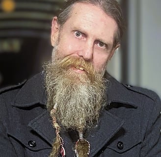 A man with a long beard is tilting their head to one side while wearing a well appointed coat