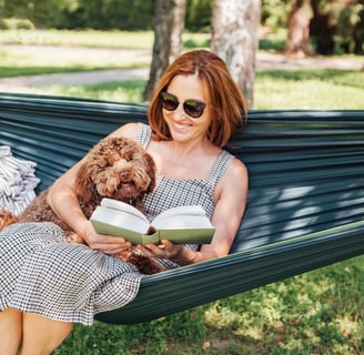 A woman relaxing in a hammock with her dog, reading a book outdoors, symbolising stress management.