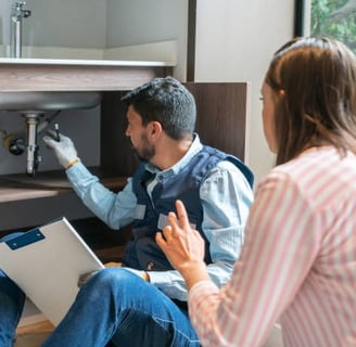 plumber pointing under a sink and explaining things to a woman