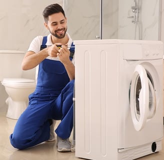 A smiling male technician is crouching down to repair the side panel of a white dryer with a screwdr