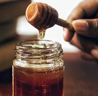 Hand holding a honey dipper dripping golden honey into a glass jar, creating a warm scene