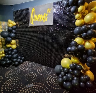 Black shimmer wall with a balloon garland wrapping both sides in black and gold with Queens sign in the middle in Brisbane ev