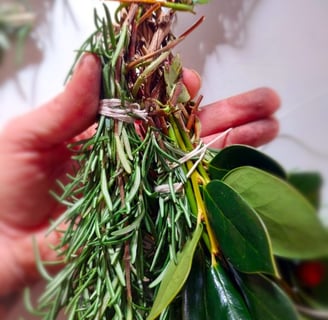 open hand holding rosemary bundle and holly onto a honeysuckle vine wreath base