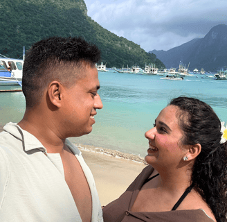 Trisha & Nikhil standing on El Nido Beach. Looking at each other with clear sea, mountains and boats