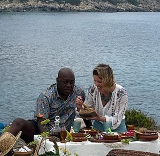 A man and woman sit on the floor, behind a low table of food, behind them is the sea.